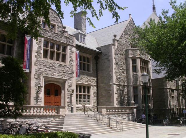 Exterior Houston Hall from Spruce Street. Grey stone building framed by trees, staircase leads to large wooden doors. Second floor windows feature blue and red Penn banners.